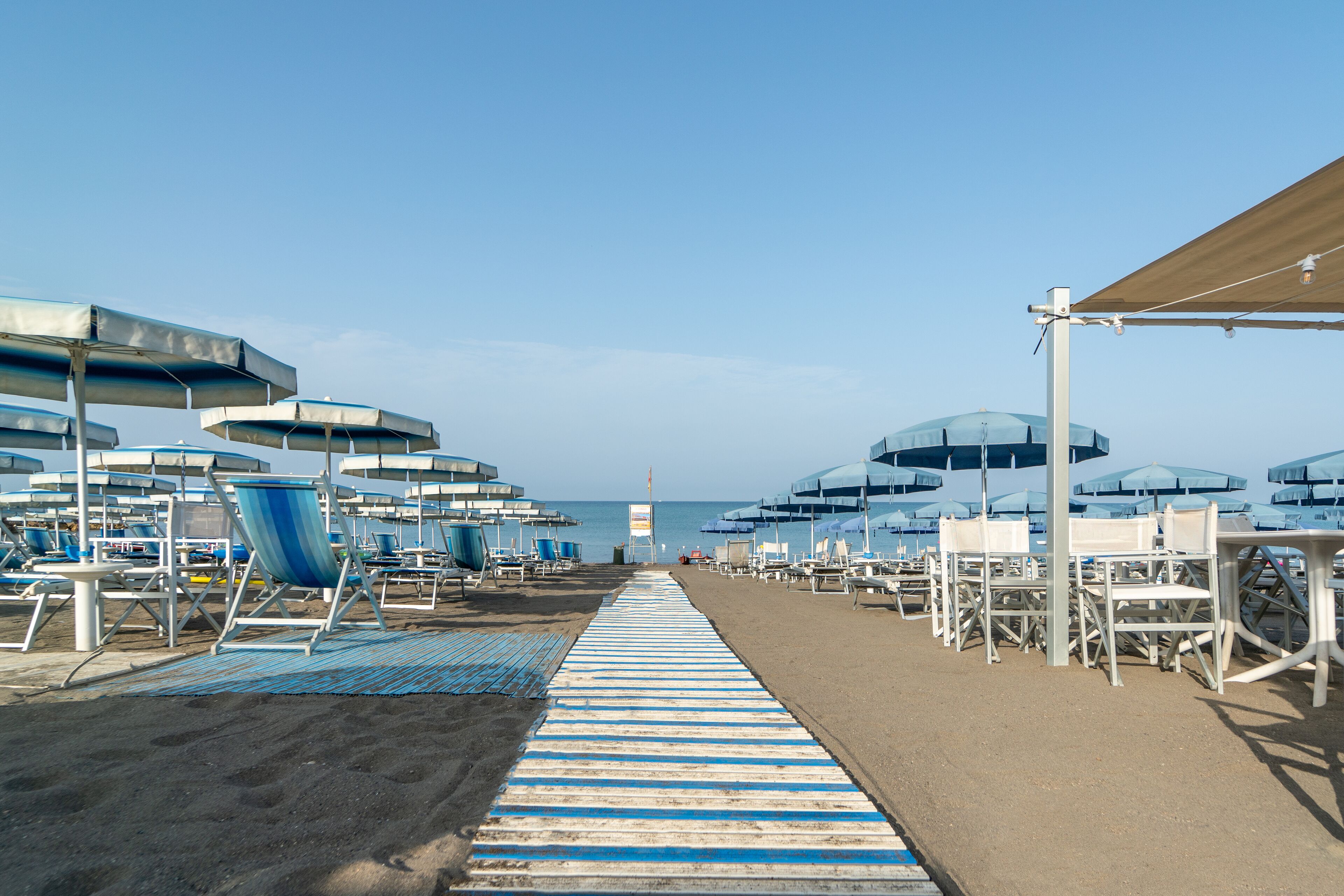 Ein Strandabschnitt mit Reihen von Liegestühlen und Sonnenschirmen, einem gestreiften Weg und Blick aufs Meer.