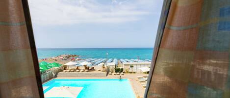 Vista de una piscina y zona de playa desde una ventana, con sombrillas y tumbonas.