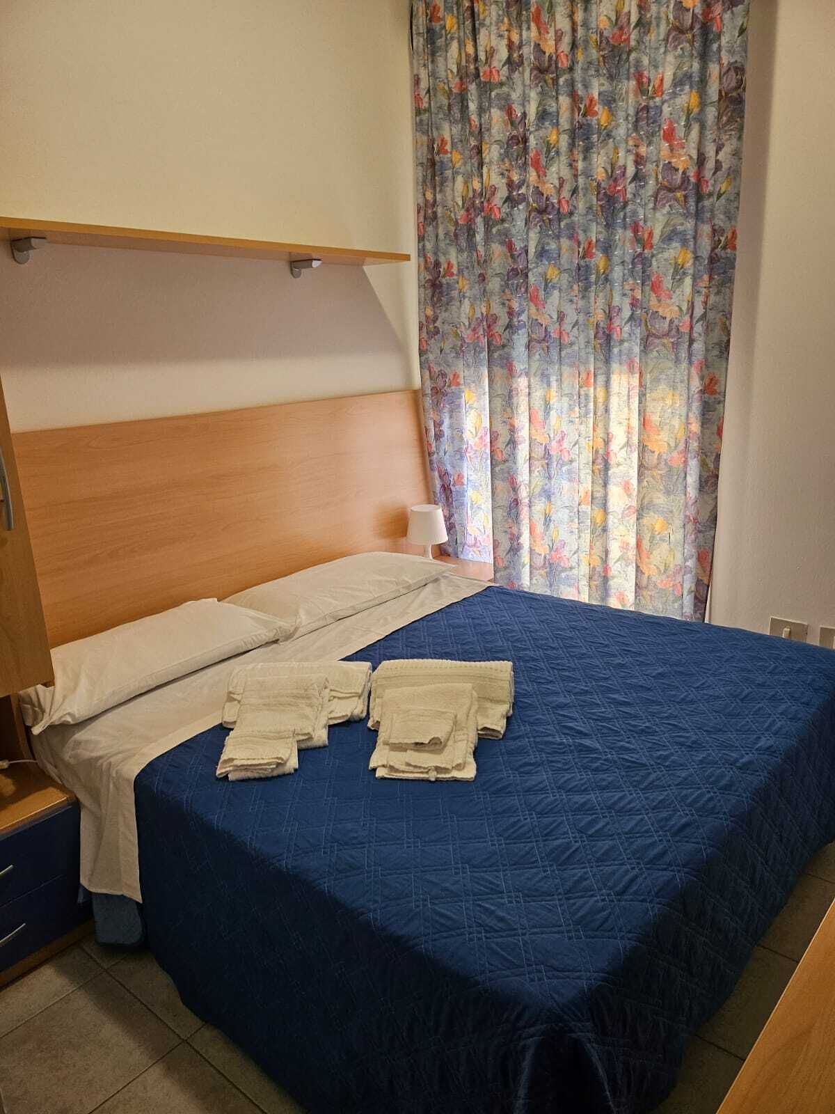 Una cama bien hecha con colcha azul, cabecera de madera y cortina estampada.