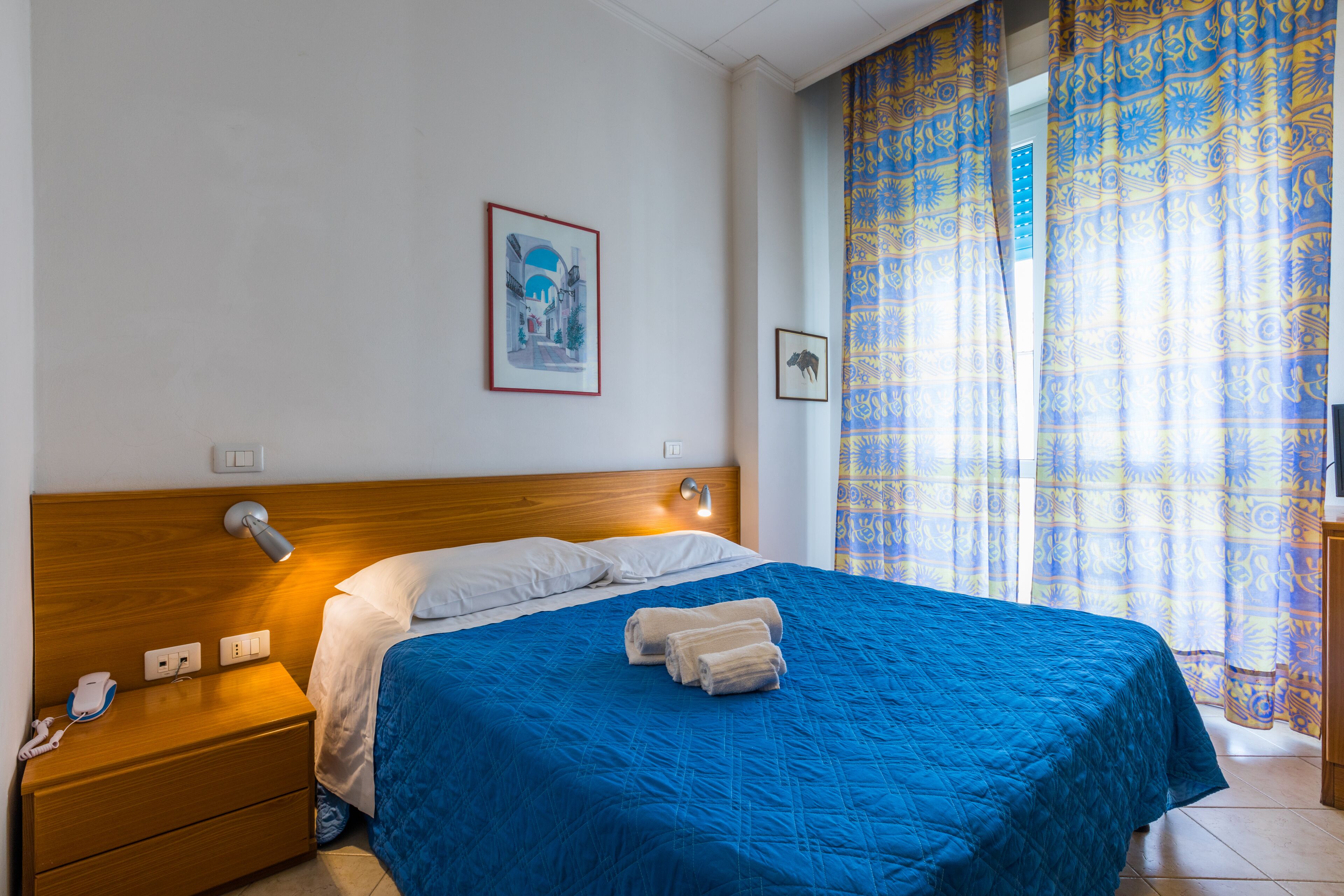 A hotel room with a wooden headboard, a blue bedspread, two rolled towels, a bedside lamp, and a framed picture on the wall.
