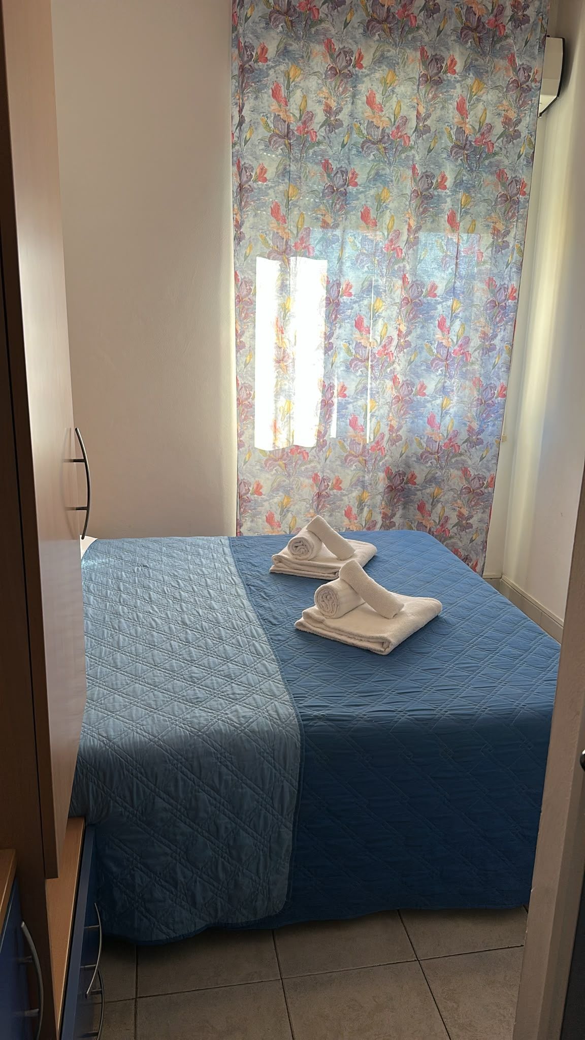 A room with a bed, a window with floral curtains, and a blue bedspread.
