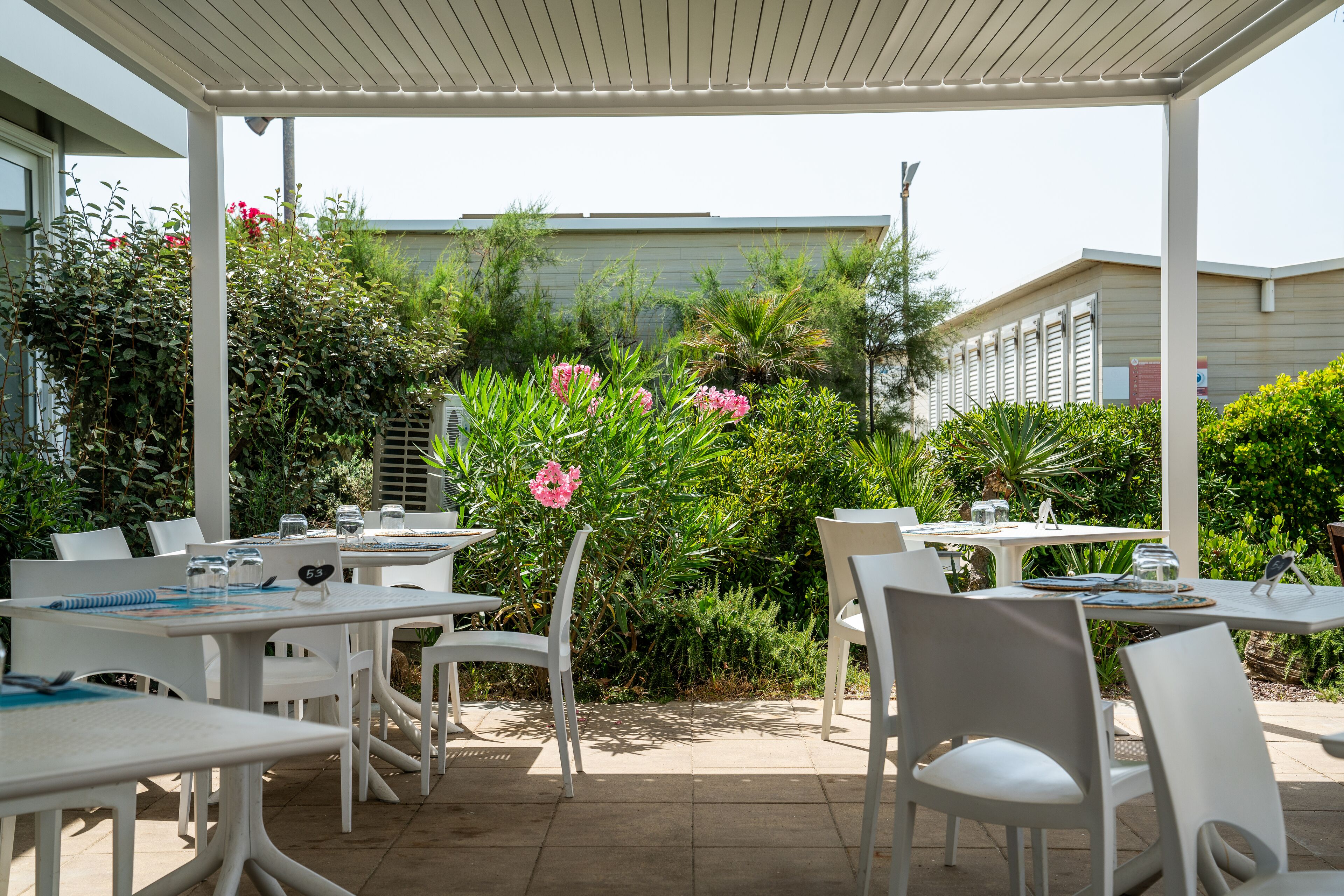 Un patio al aire libre con mesas y sillas blancas, rodeado de exuberante follaje y flores rosadas.