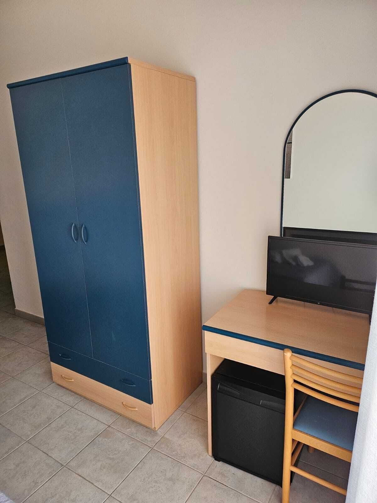 A room with a large wardrobe, a desk with a chair, a television, and a small black cabinet.