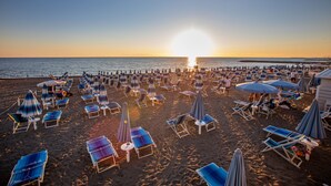 A beach with rows of sun loungers and umbrellas, a clear sky, and the sun setting over the water. - Hotel Stella Marina (Cecina)