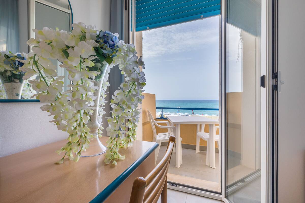Un balcón con vistas al océano, una mesa de madera con un jarrón de flores y sillas blancas.