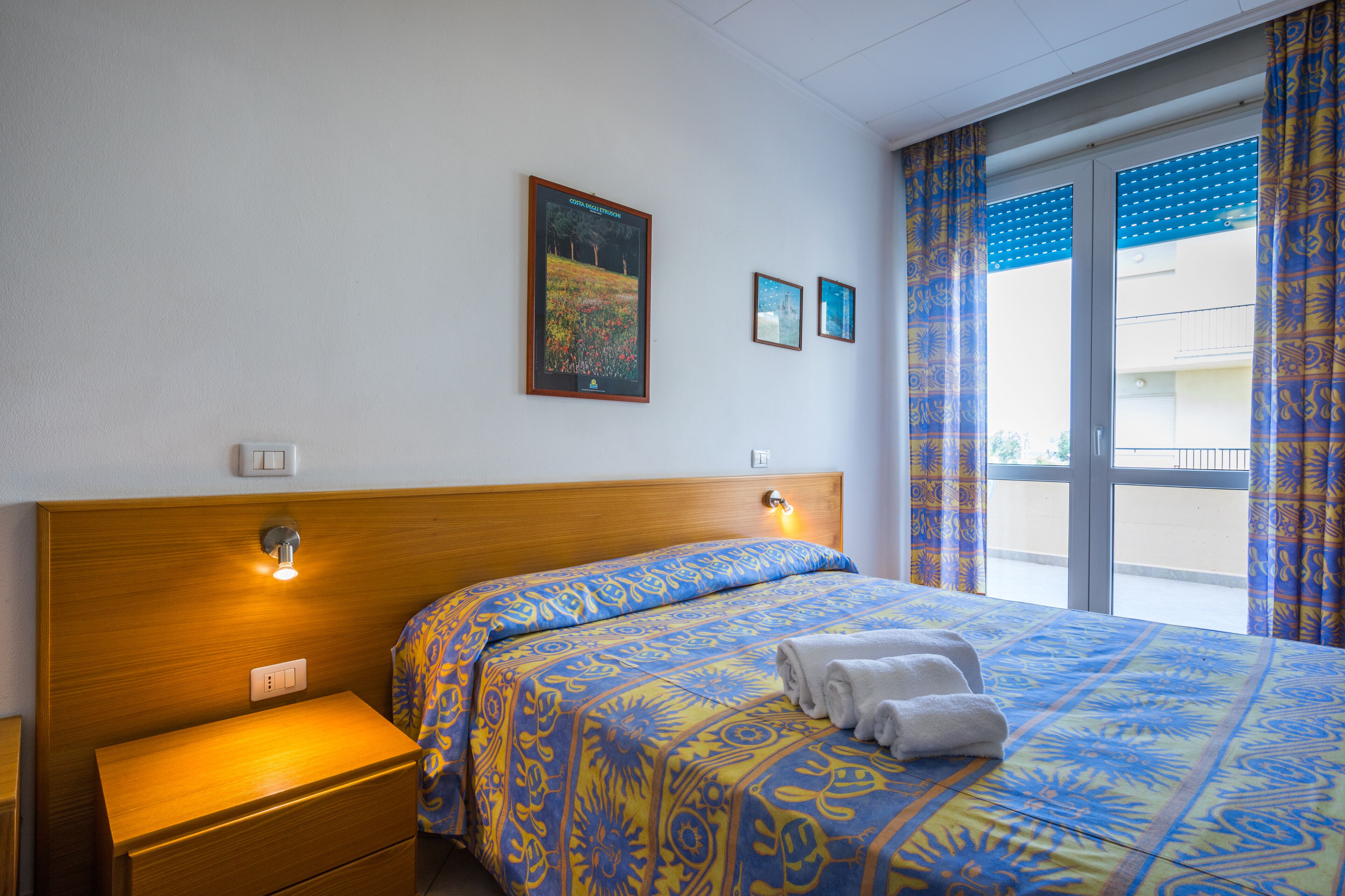 A hotel room with a wooden headboard, a bed with patterned bedding, a nightstand, and a window with curtains.