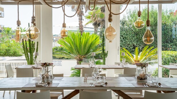 A restaurant with tables set for dining, hanging light fixtures, and a view of greenery outside. - Hotel Stella Marina (Cecina)