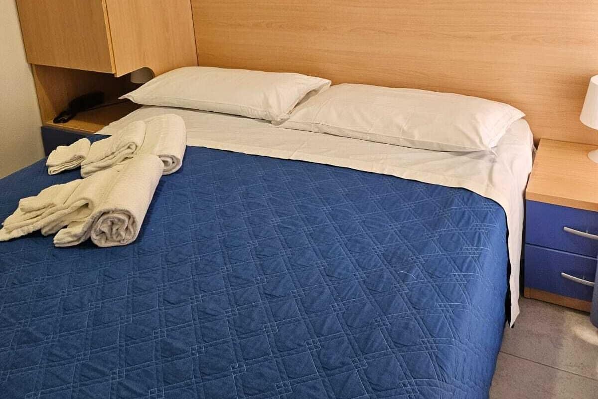 Una cama bien hecha con colcha azul, almohadas blancas y toallas enrolladas sobre un cabecero de madera.