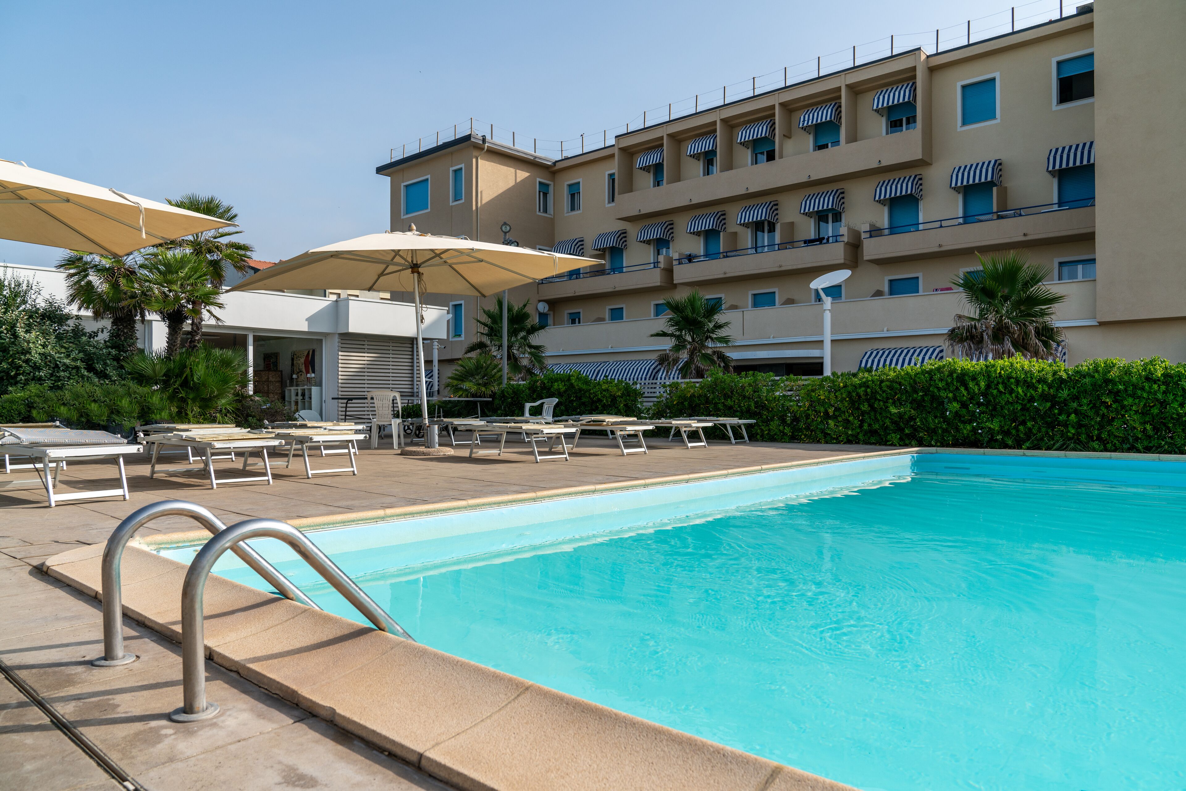 A swimming pool with lounge chairs and umbrellas, surrounded by a building with balconies and striped awnings.