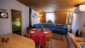 Ein gemütliches Wohnzimmer mit Holzbalkendecke, einem blauen Sofa, einem Esstisch mit roter Tischdecke und einer Küchenzeile mit Herd.