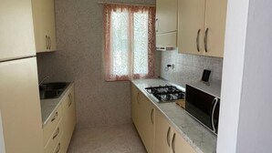 A compact kitchen with beige cabinets, a gas stove, and a window with patterned curtains. - Spacious Apartment With Terrace in Rosolina Mare (Rosolina)
