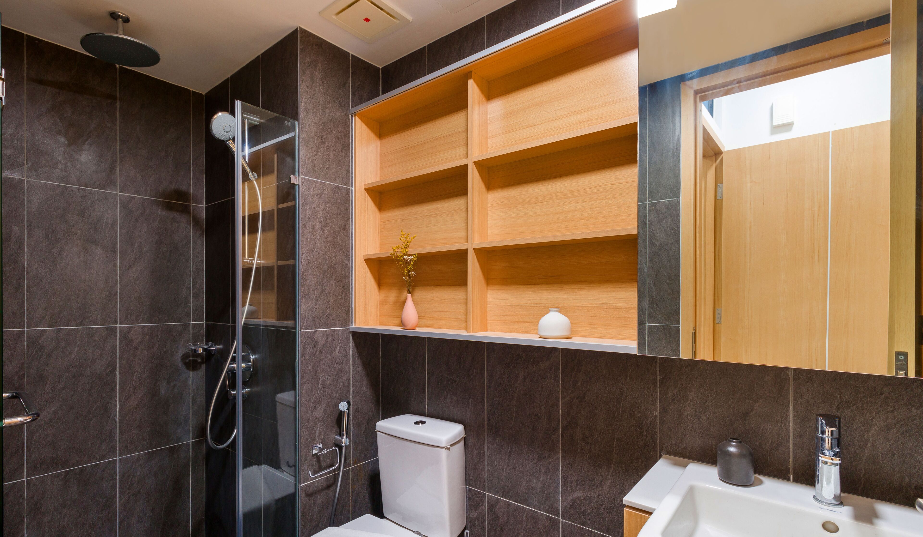 A modern bathroom with dark tiles, a glass-enclosed shower, a wooden vanity with a mirror, and a toilet.