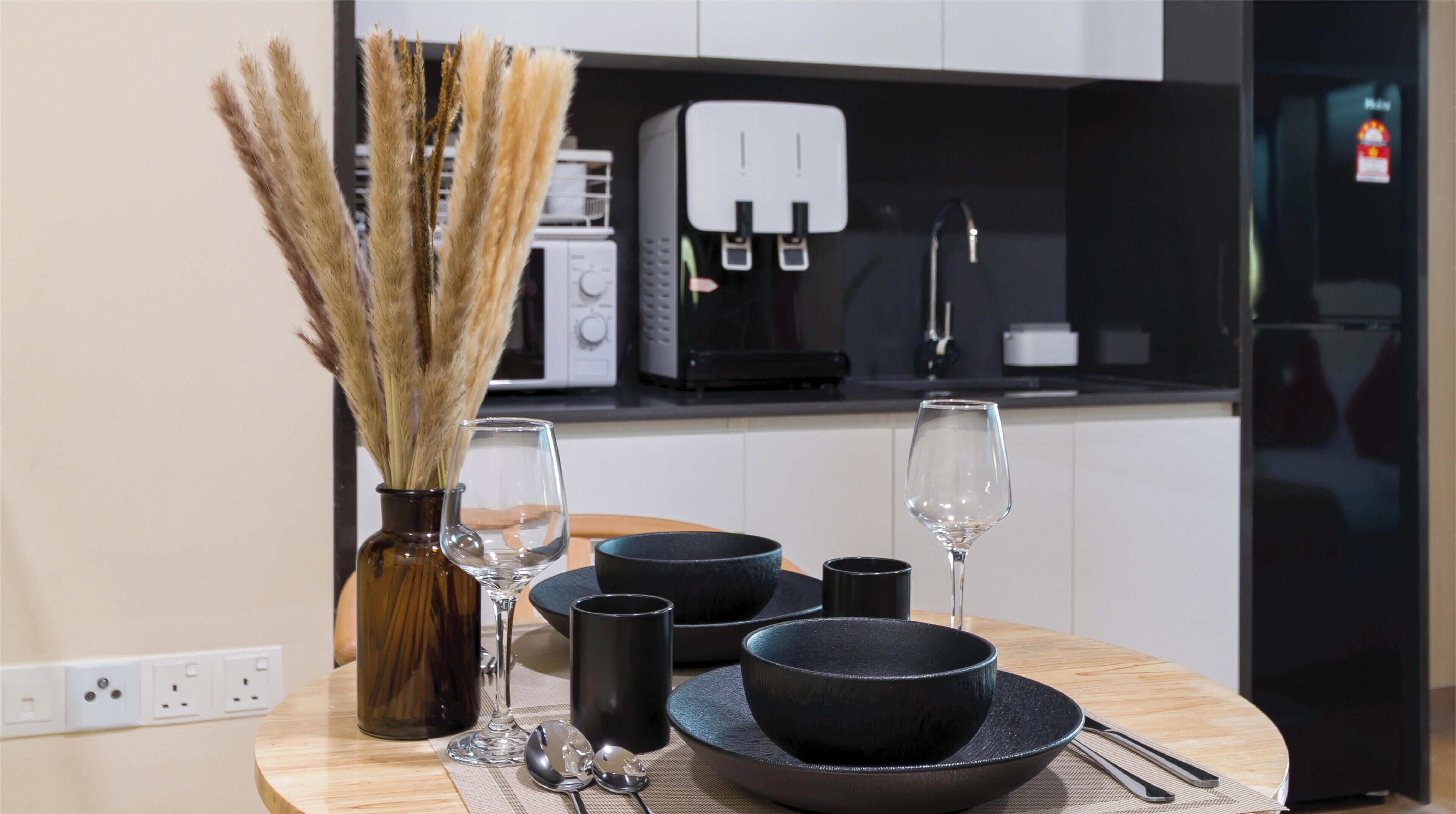 A modern kitchen with a round wooden table set for dining, featuring black bowls and glasses, and a vase with dried plants.