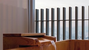 Um banco de madeira com um elemento de água, vista da cidade através de uma janela com persianas verticais e o logotipo "FIVE SENSES EXPERIENCE SUITE".