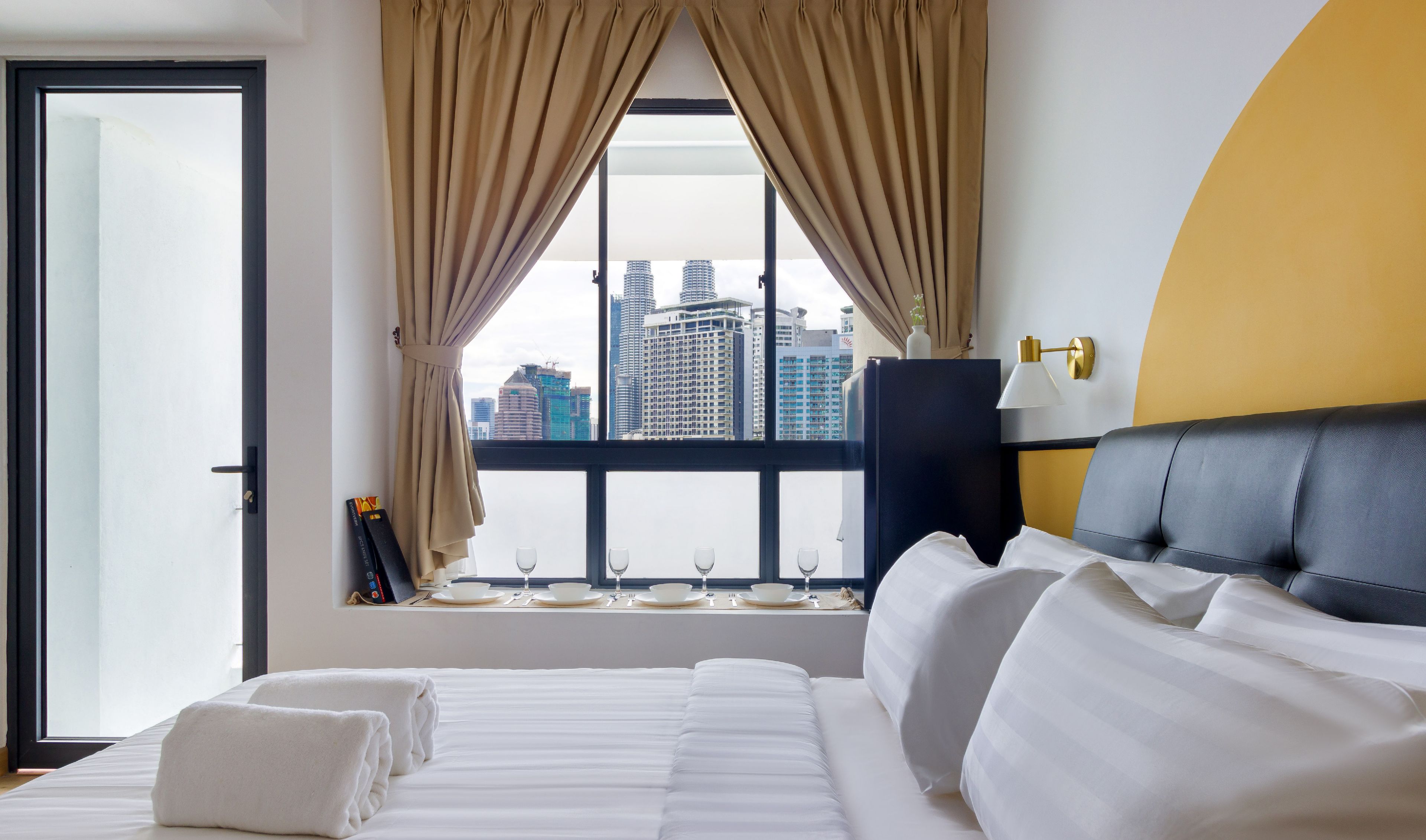 A hotel room with a large bed, a view of the city through a window, and a yellow accent wall.