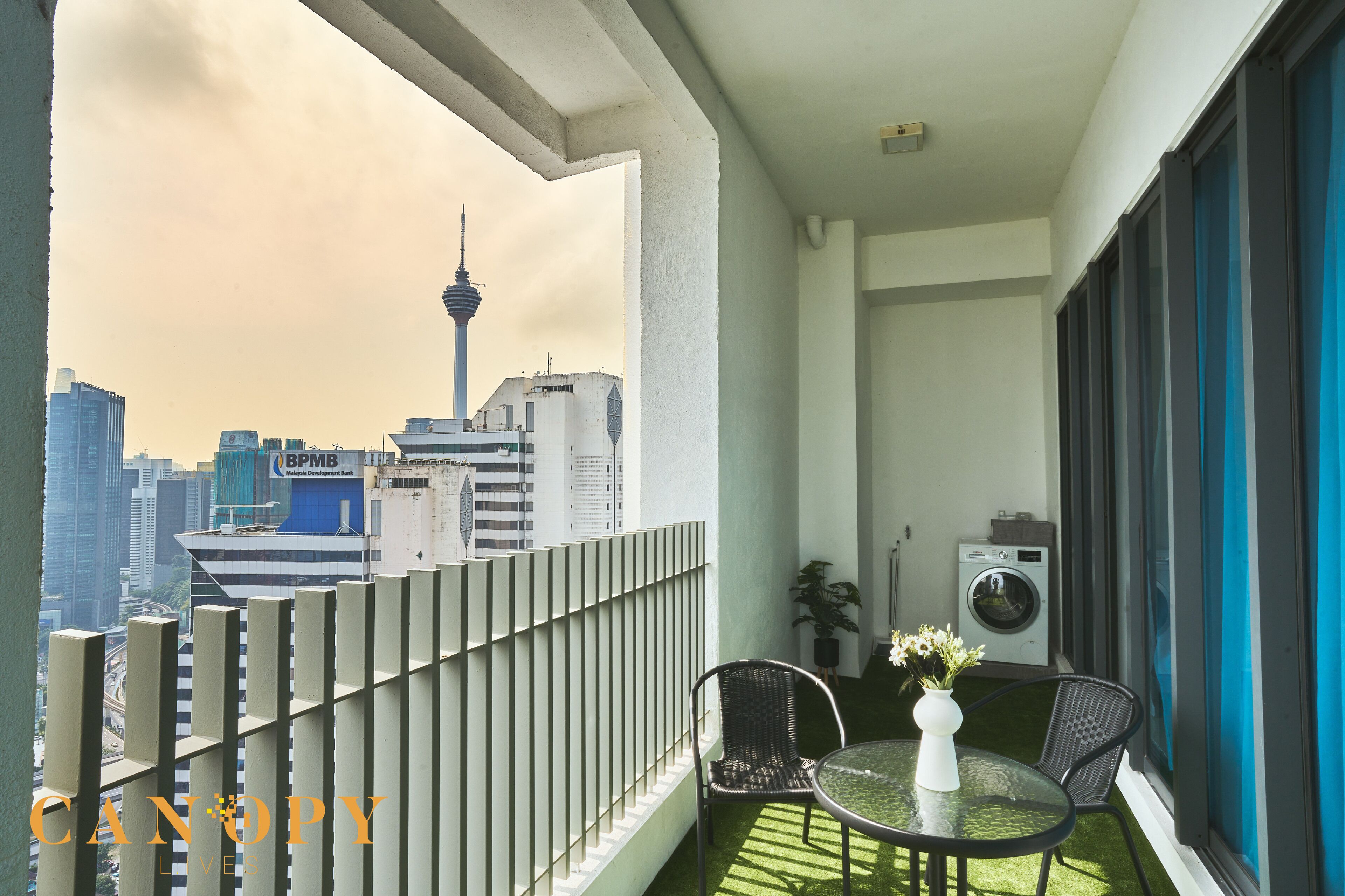 Een balkon met uitzicht op een stadshemelbeeld, inclusief een toren en een gebouw met een blauw bord. Er staat een klein tafeltje met een bloevas en twee stoelen.
