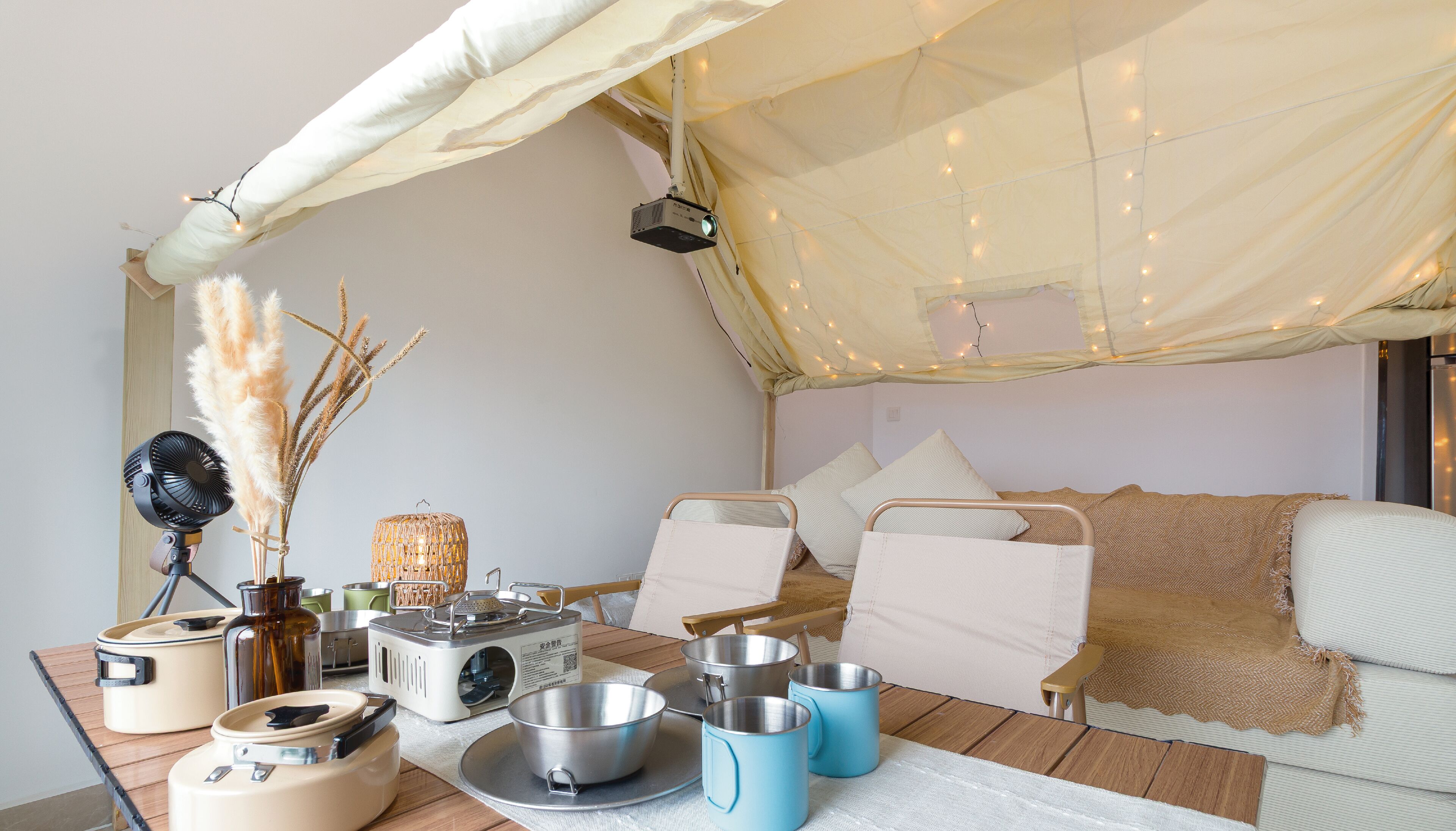 Une pièce confortable, bien éclairée et aménagée comme une tente, avec un lit, une table équipée d’ustensiles de cuisine et un ventilateur.