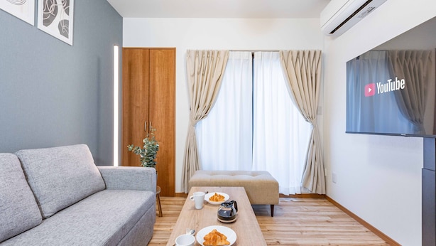 A modern living room with a grey sofa, a wooden coffee table with plates of food, a small table lamp, and a TV displaying the YouTube logo. - Fleur Sanno (Osaka)