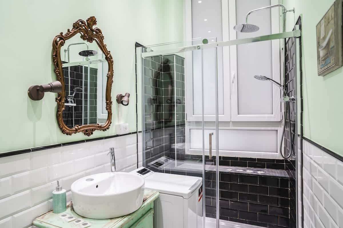 Une salle de bain avec un lavabo blanc, un miroir doté d’un cadre orné, une douche avec une paroi en verre et un sol carrelé.