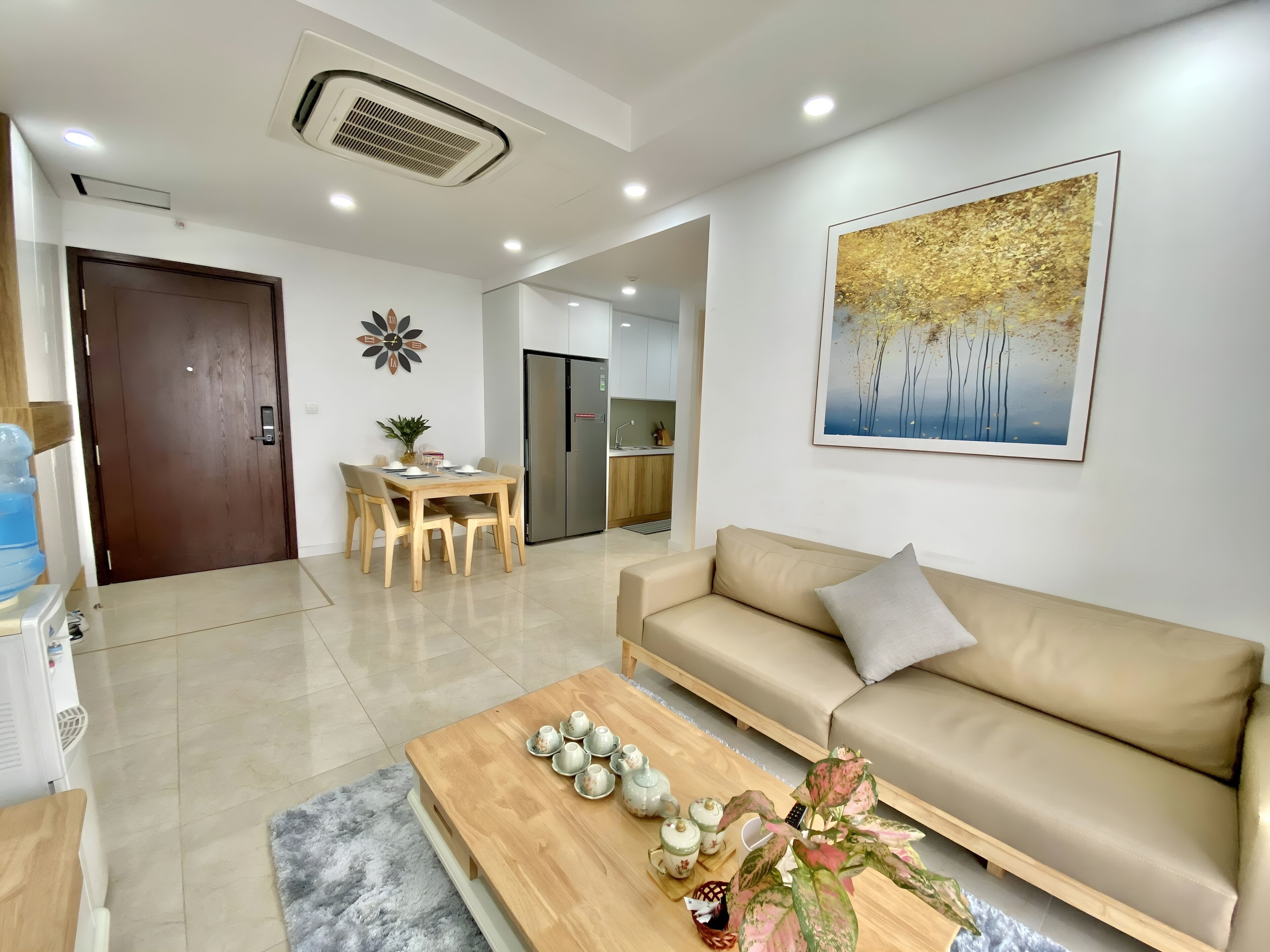 A modern living room with a beige sofa, a wooden coffee table with teapots, a dining area with a table and chairs, and a kitchen in the background.