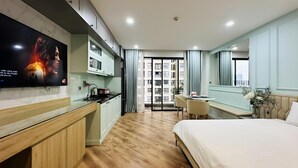 A modern hotel room with a wooden floor, a bed, a TV, a kitchen area, and a balcony with curtains. - Vinhomes D'Capitale Ha Noi-Gem Apartment (Hanoi)