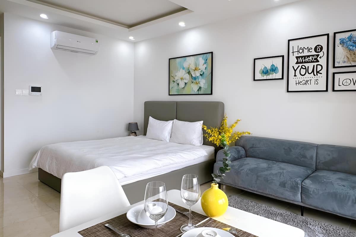 A modern hotel room with a bed, a gray sofa, a dining table with a vase of yellow flowers, and wall art.