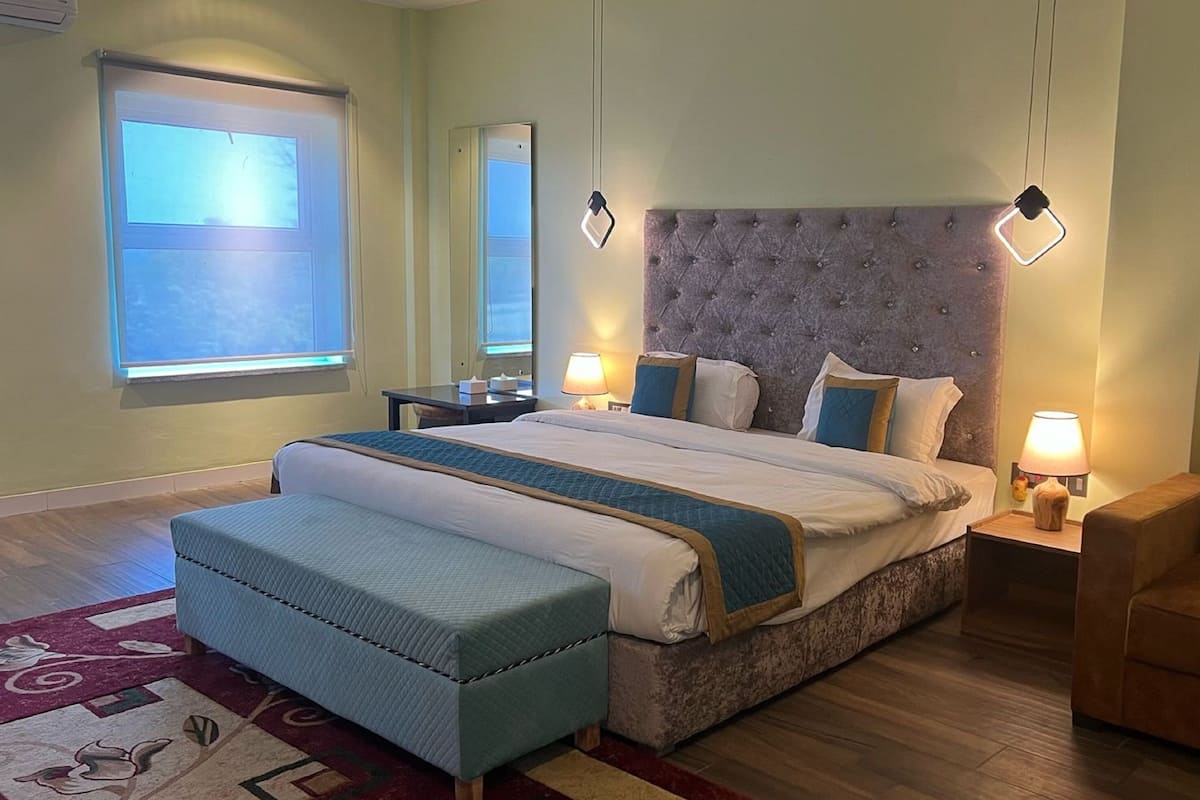 Une chambre d’hôtel avec un grand lit, un coin salon avec un fauteuil, une table de chevet, un miroir et une fenêtre avec des stores.