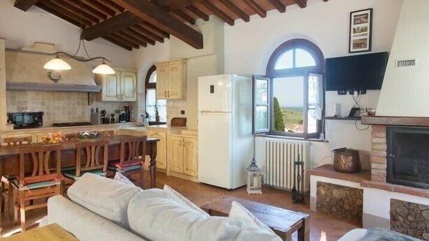 A spacious living area with a wooden ceiling, a stone fireplace, a large window, and a kitchen with wooden cabinets and a white refrigerator. - Rustikale Villa mit Privat Pool in Buti, Toscana. Haustierfreundlich (Buti)