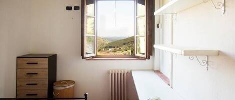 A bedroom with a bed, a desk, a radiator, a window with an open shutter, and a view of the countryside.