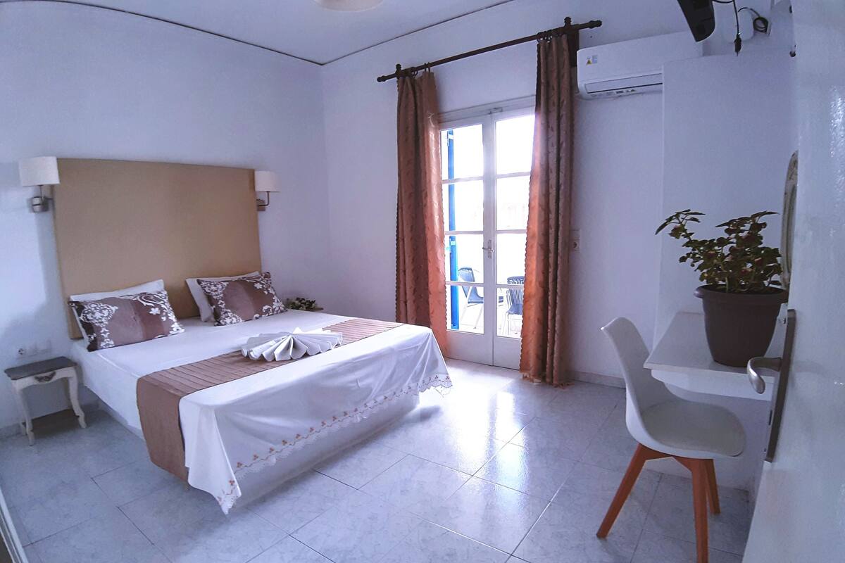 A hotel room with a bed, bedside table, chair, potted plant, and a view of the outside.