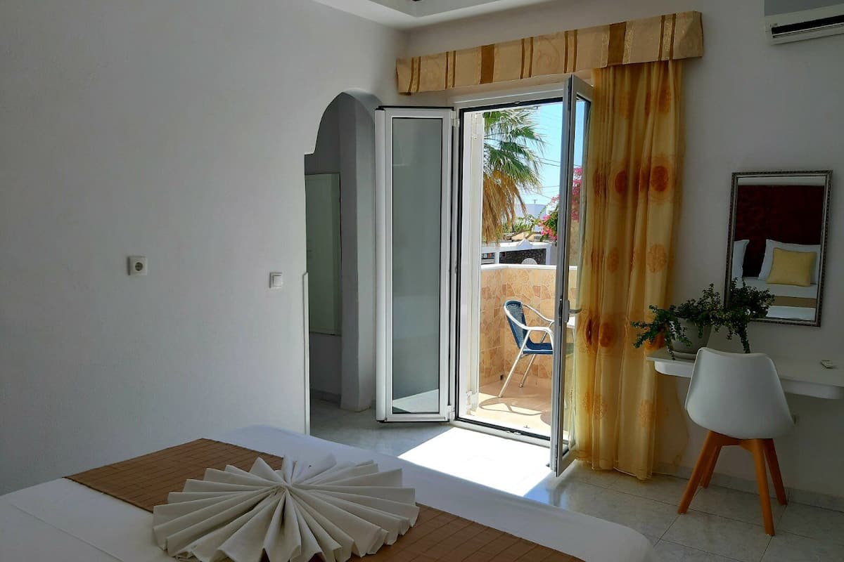 A hotel room with a bed, a large window with curtains, a sliding glass door leading to an outdoor area, and a small white chair.