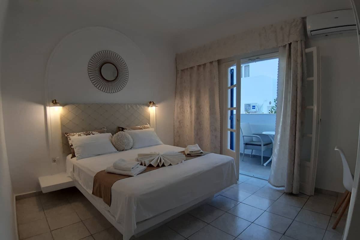 A neatly arranged bedroom with a bed, bedside tables, a wall-mounted mirror, and a view of the balcony through an open door.