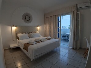 A neatly arranged bedroom with a bed, bedside tables, a wall-mounted mirror, and a view of the balcony through an open door. - Summer Holiday by Atalos Suites (Santorini)