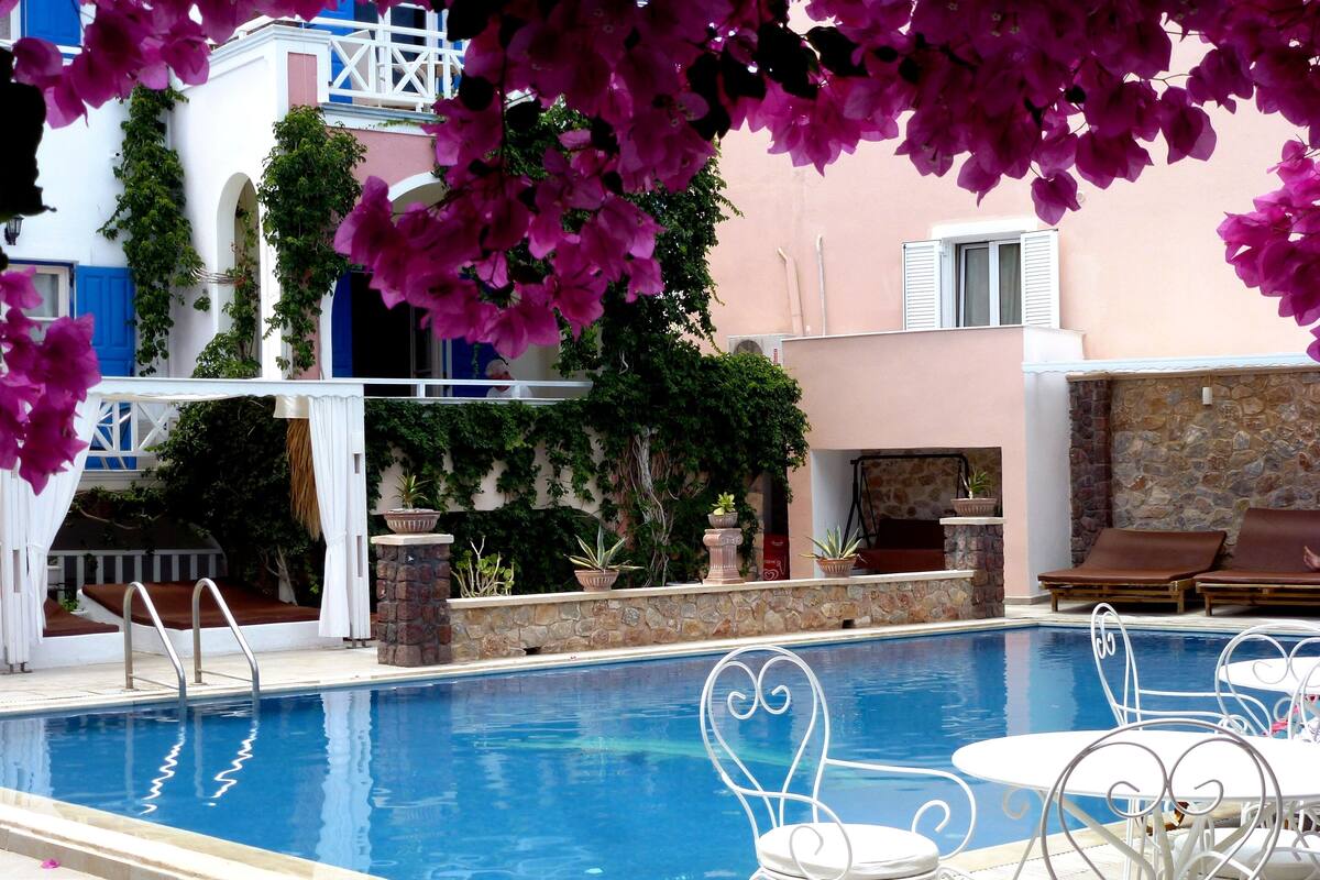 A swimming pool with white chairs and tables, surrounded by potted plants and a stone wall.