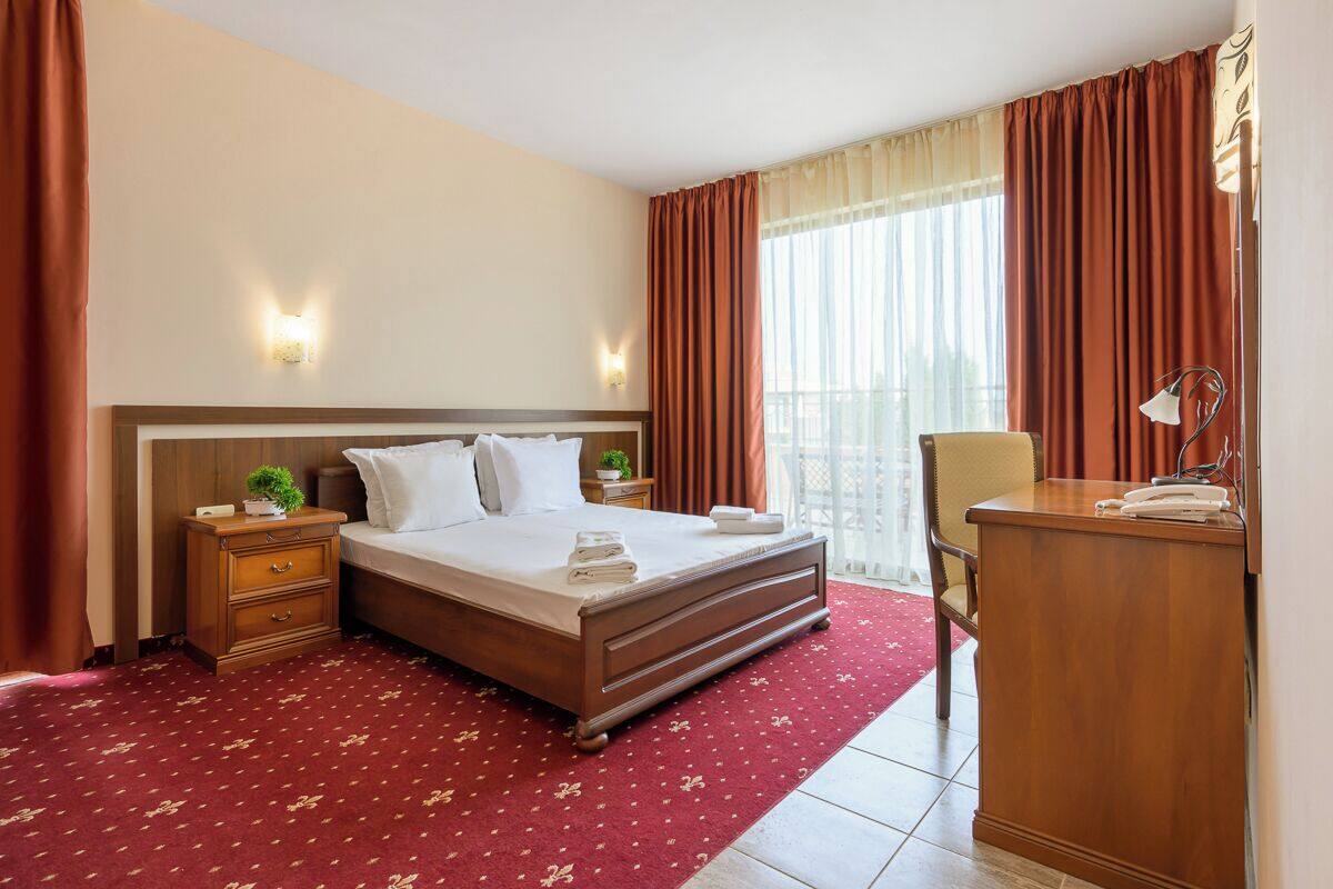 A hotel room with a large bed, a wooden nightstand with a lamp, a desk with a telephone, and a view of the outdoors through a window with curtains.