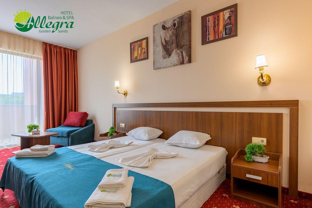 Een hotelkamer met een groot bed, twee nachtkastjes, een rode stoel, een kleine tafel met een plant en kunstwerken aan de muren.
