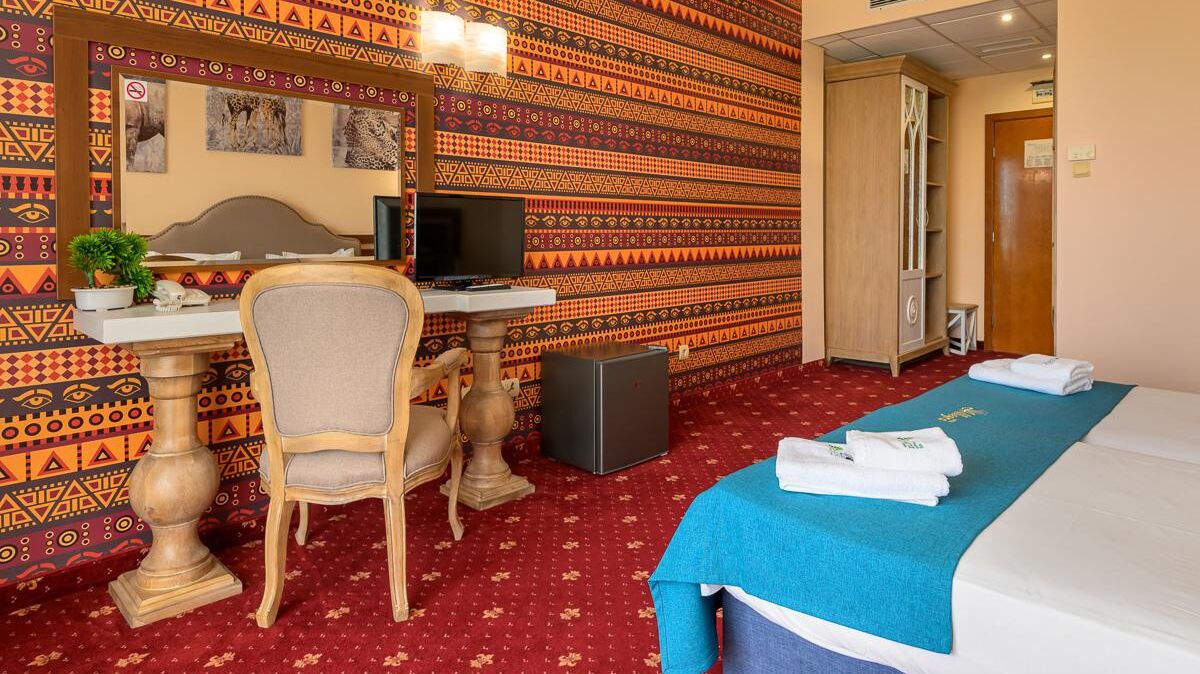 A hotel room with a bed, a desk with a computer, a chair, a small table with a plant, and a wardrobe.