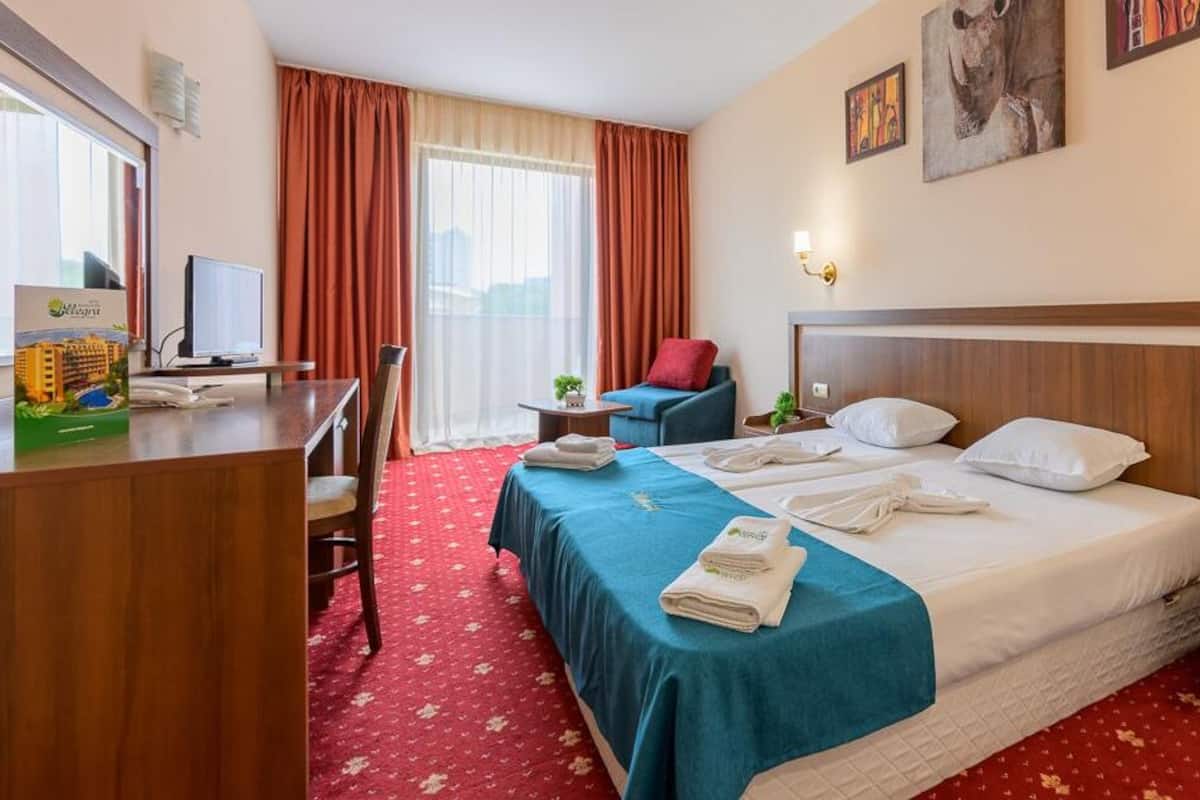 A hotel room with a large bed, a desk with a computer, a chair, a small table with a plant, and a window with curtains.