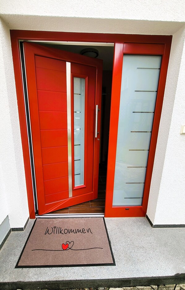 A red door with a glass panel and a doormat with "Willkommen" and a heart symbol.