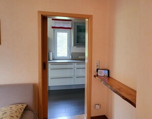 Um corredor estreito leva a uma cozinha com armários brancos, uma janela com um valance vermelho, micro-ondas embutido e termostato na parede.