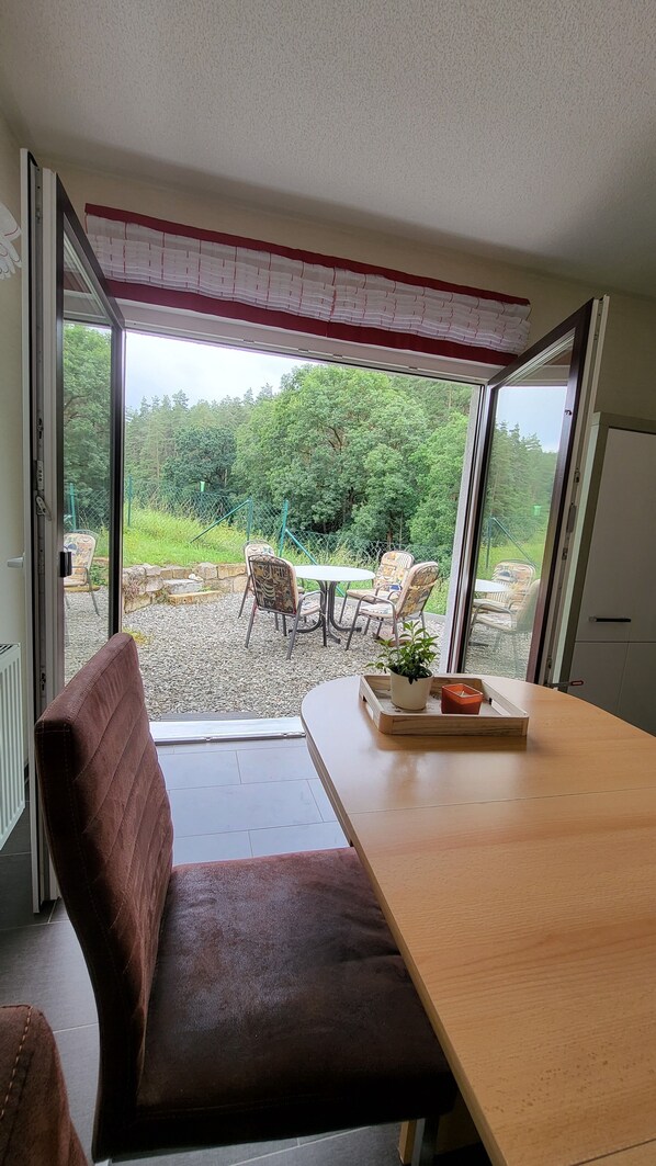 Une pièce avec une table en bois, un banc marron avec un coussin, et une vue sur un espace extérieur aménagé avec une table et des chaises.
