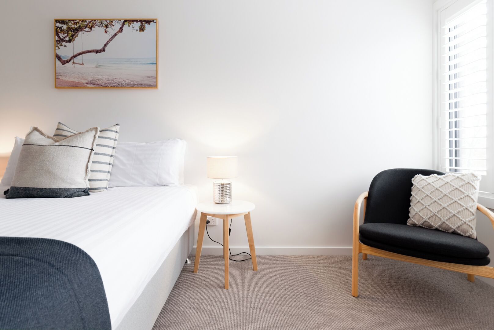 A hotel room with a bed, bedside table with a lamp, a chair with a cushion, and a wall-mounted picture.