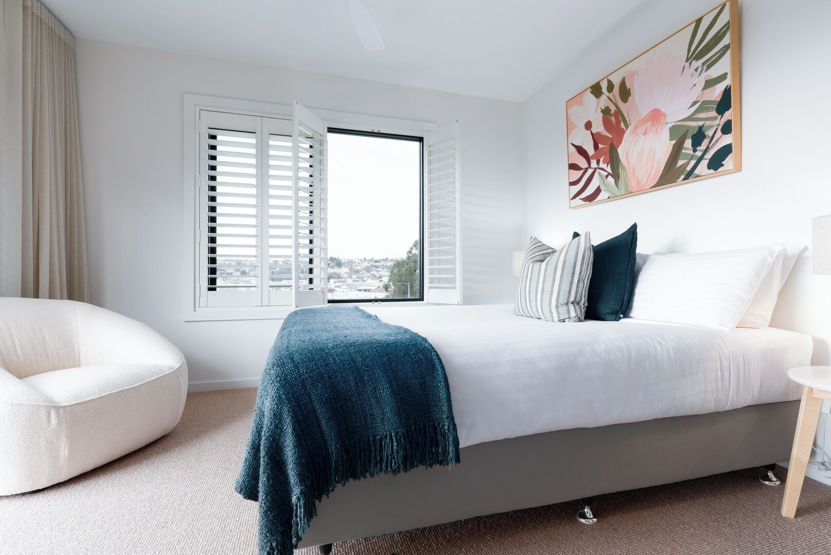 A modern bedroom with a bed, a chair, a window with shutters, and a painting on the wall.