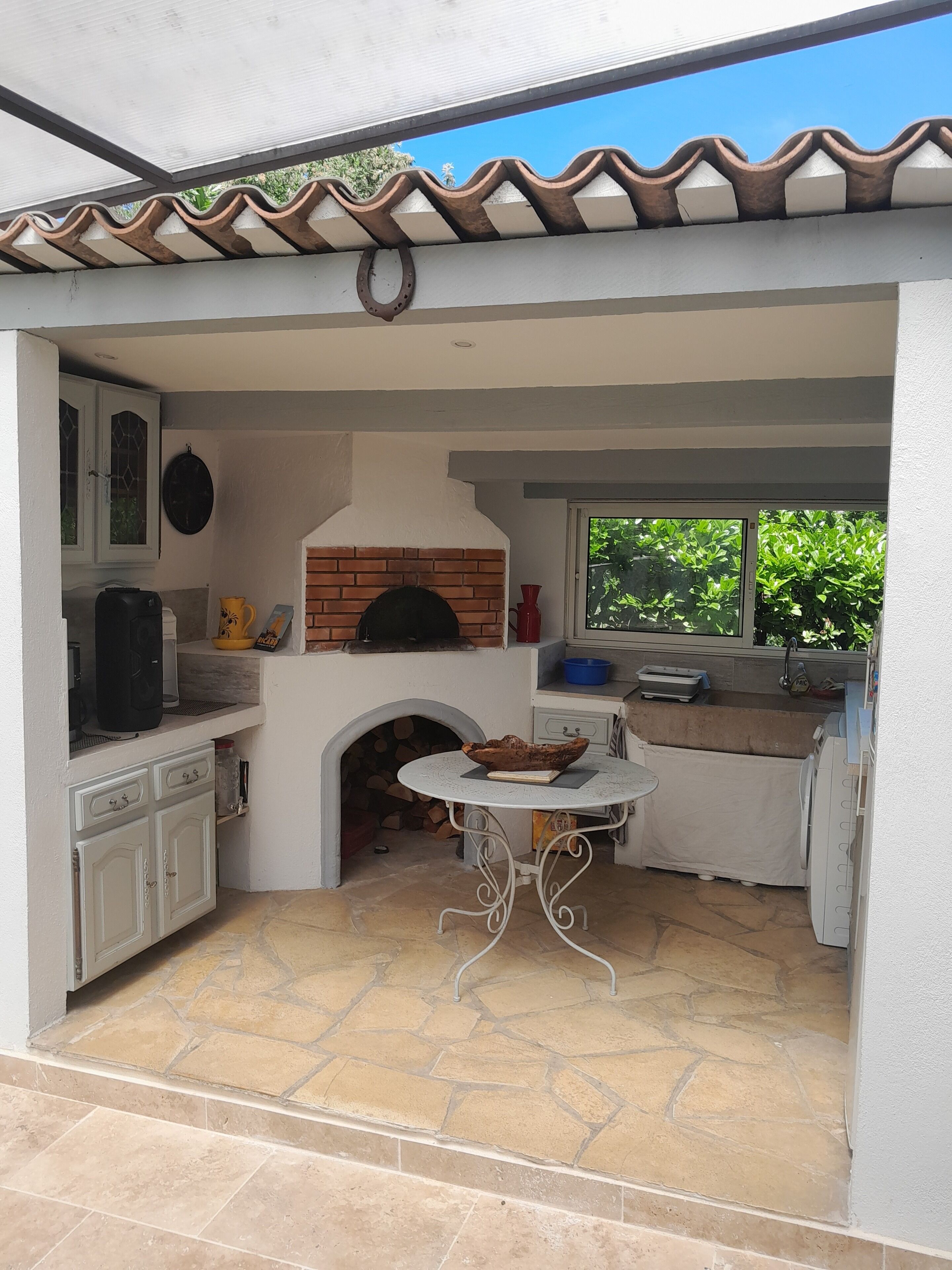 Un espace cuisine extérieur doté d’un four en briques, de meubles blancs et d’un sol carrelé en pierre.