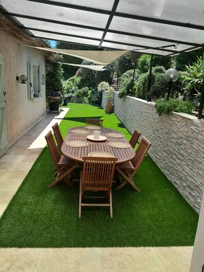 Eine Terrasse mit einem runden Holztisch und Stühlen, umgeben von künstlichem Grün, unter einem überdachten Bereich mit einem Metalldach.