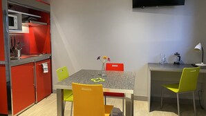 A compact kitchen with a built-in stove, a dining table with chairs, a TV mounted on the wall, and a small workspace with a lamp.