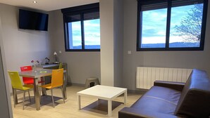 A modern living room with a brown leather sofa, a dining table with colorful chairs, a TV mounted on the wall, and large windows with a view of trees.