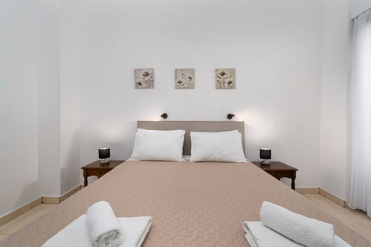 Un dormitorio ordenado con una cama grande, dos mesitas de noche con lámparas y tres cuadros florales colgados en la pared.