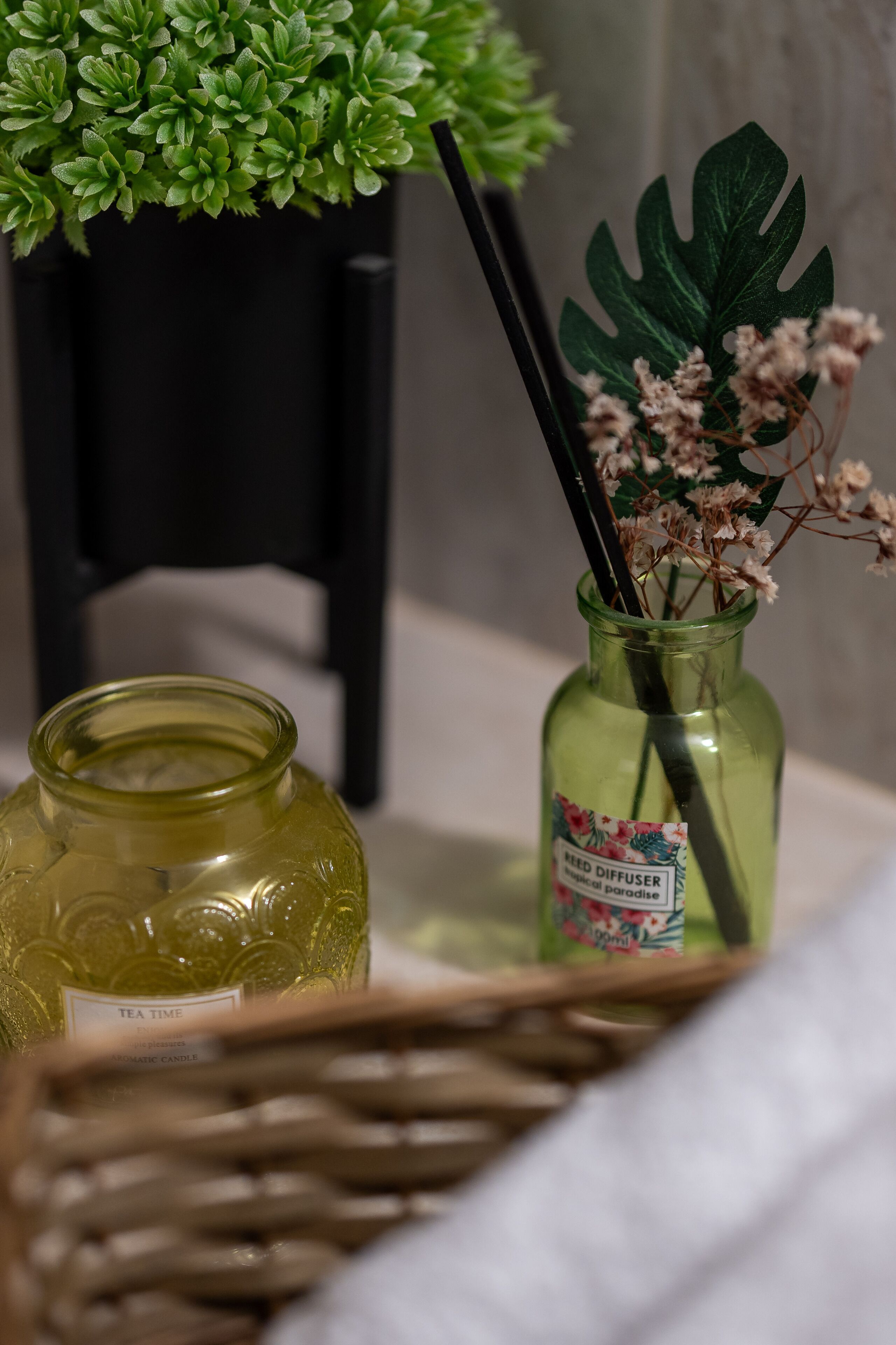 Eine grüne Glasflasche mit Etikett, ein schwarzer Stuhl und ein Korbwicker-Korb mit einer grünen Pflanze sowie einer Vase mit Blumen.