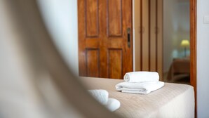 Ein ordentlich bezogenes Bett mit einem gefalteten weißen Handtuch, eine Holztür im Hintergrund und ein Blick in einen Raum mit einem Bett und einer Lampe.